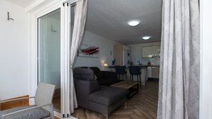 Ein modernes Wohnzimmer mit einer Couch, einem kleinen Tisch und einem Sessel. Im Hintergrund ist eine Küchenzeile mit einer Arbeitsplatte und Schränken zu sehen.