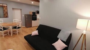 Ein Wohnzimmer mit einer schwarzen Couch, zwei weißen Sesseln, einem kleinen runden Tisch und einer Küchenzeile im Hintergrund.
