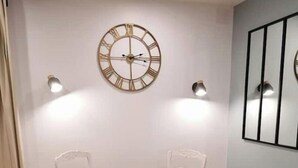 Ein Essbereich mit einem runden Holztisch und vier weißen Stühlen mit schwarzen Kissen. Darüber an der Wand hängt eine große Wanduhr mit römischen Ziffern.