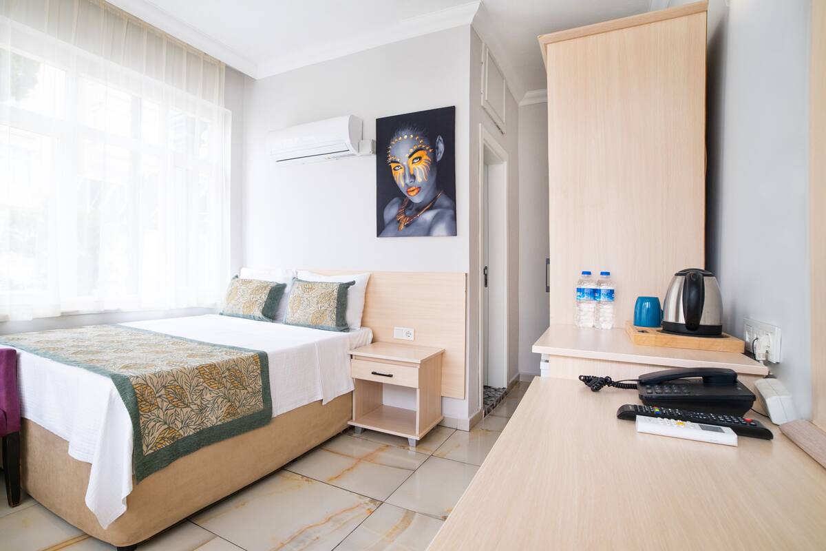 Habitación de hotel con cama, un sillón morado, escritorio con hervidor y un cuadro en la pared.