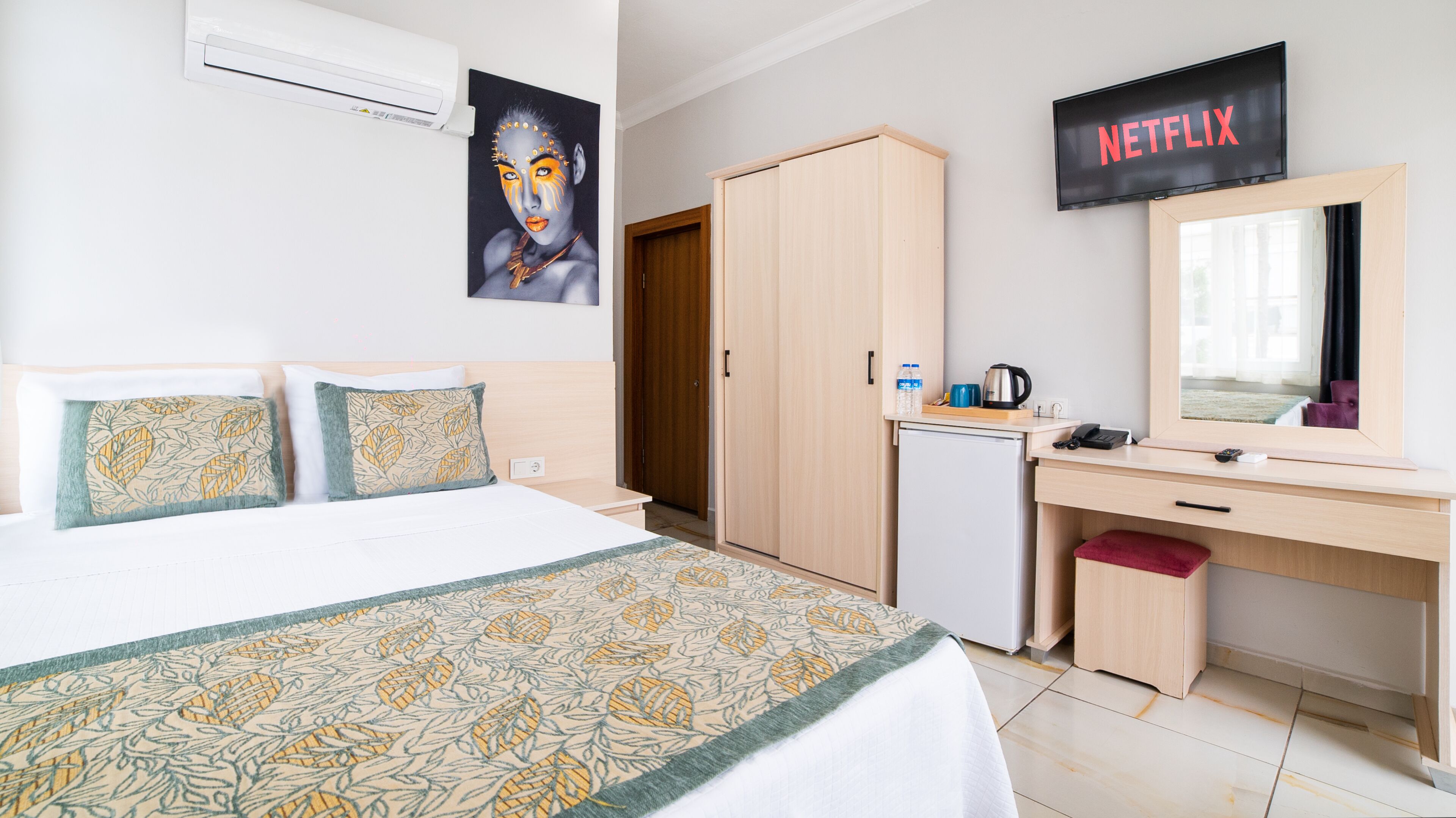 A hotel room with a bed, a desk, a wardrobe, a mirror, a TV, and a small fridge.