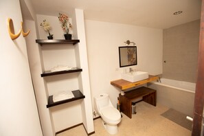 Une salle de bain moderne équipée de toilettes, d’un lavabo et d’une baignoire. On y trouve des étagères en bois où sont pliés des serviettes, ainsi que des plantes en pot.