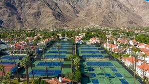 Ein Tennisplatzkomplex mit mehreren Plätzen, umgeben von Wohngebäuden und Palmen, mit Bergen im Hintergrund.