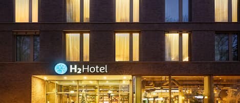 A hotel entrance with a glass facade, a sign reading "H2Hotel", and a visible reception area inside.