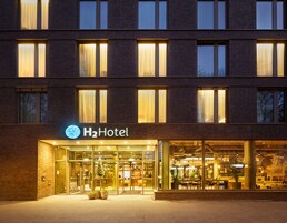 Entrada de hotel con fachada de vidrio, un cartel que dice "H2Hotel" y un área de recepción visible en el interior.