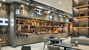 A modern cafe with a large sign displaying the name 'eH+Mo+He+H'. It features a counter with various food items, shelves with plants, and seating areas with tables and chairs. - H2 Hotel Mainz (Mainz)