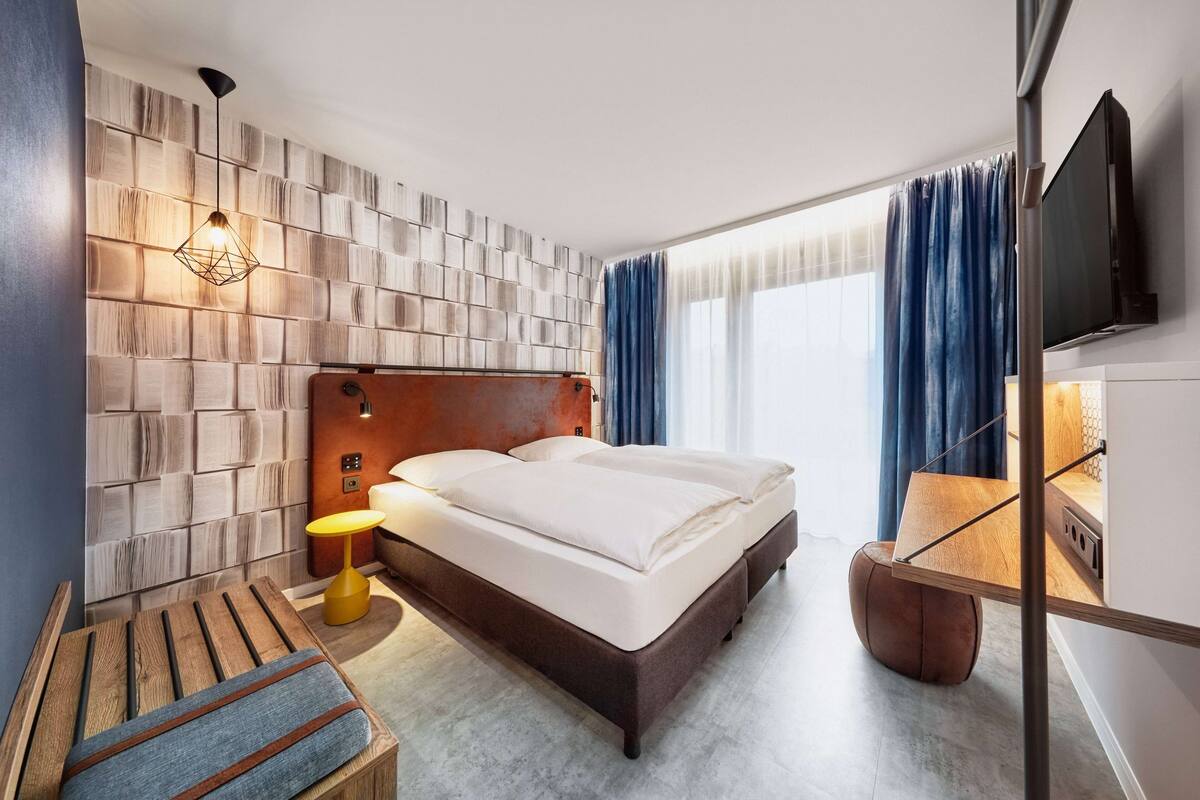 A hotel room with a bed, a wooden bench, a table with a lamp, and a TV mounted on the wall.