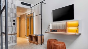 A modern hotel room with a large flat-screen TV mounted on a wall, a wooden shelf with a decorative item, and two leather ottomans on the floor. - H2 Hotel Mainz (Mainz)