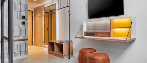 Quarto de hotel moderno com uma grande TV de tela plana na parede, uma prateleira de madeira com um objeto decorativo e dois pufes de couro no chão.