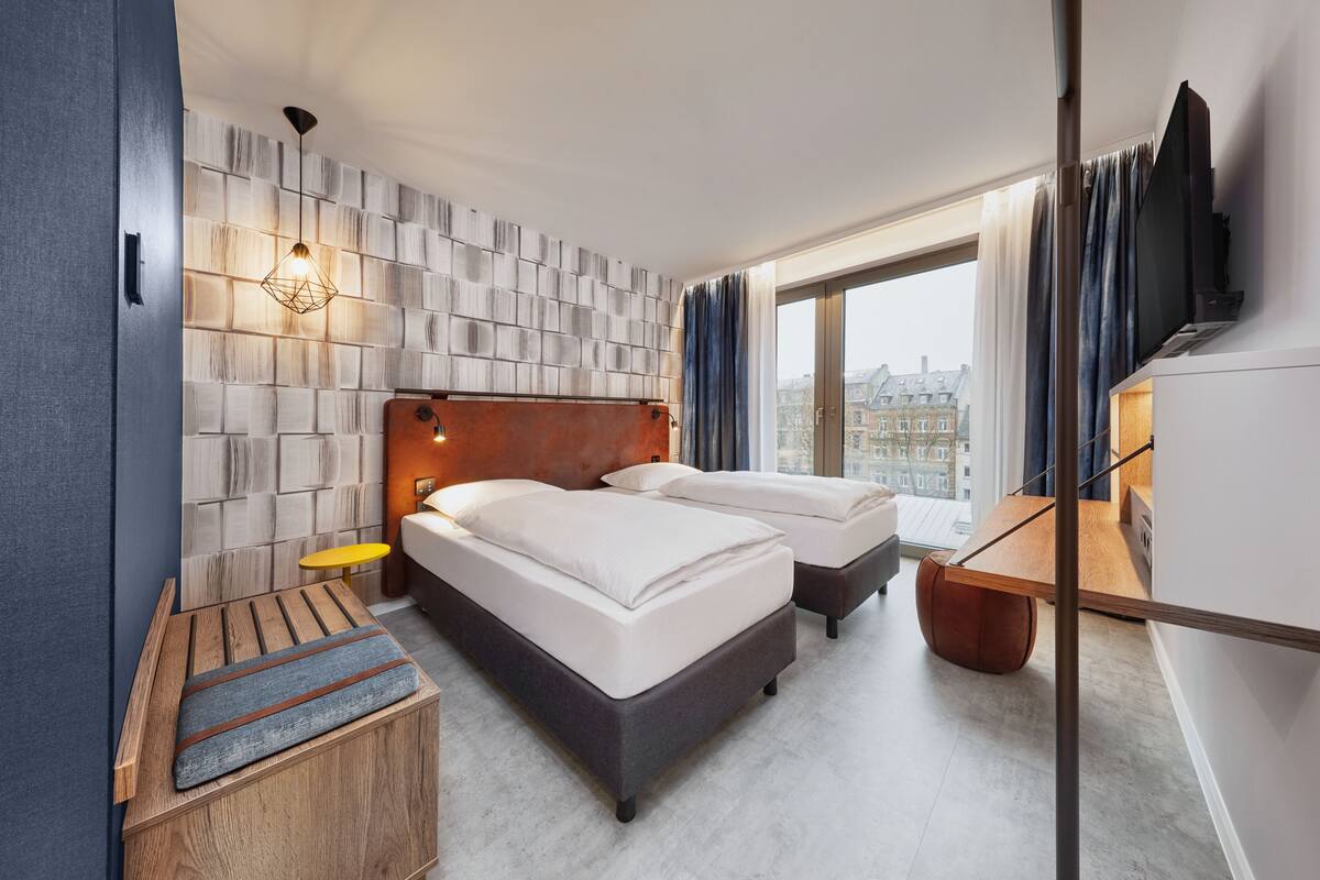A hotel room with two beds, a wooden bench, a small table, a wall-mounted TV, and a large window with curtains.