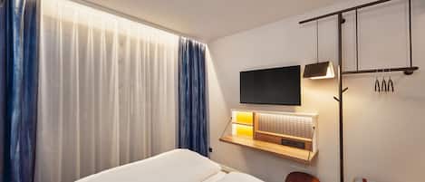 A hotel room with a bed, a TV mounted on the wall, a bedside table with a lamp, and a window with curtains.