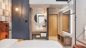 A modern hotel room with a large bed, a wooden headboard, a round mirror, a built-in sink, and a wooden bench. - H2 Hotel Mainz (Mainz)