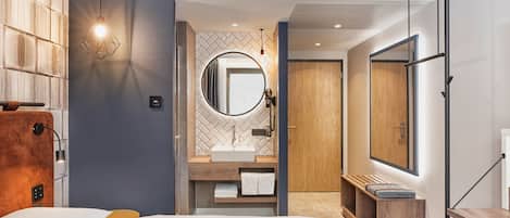 A modern hotel room with a large bed, a wooden headboard, a round mirror, a built-in sink, and a wooden bench.