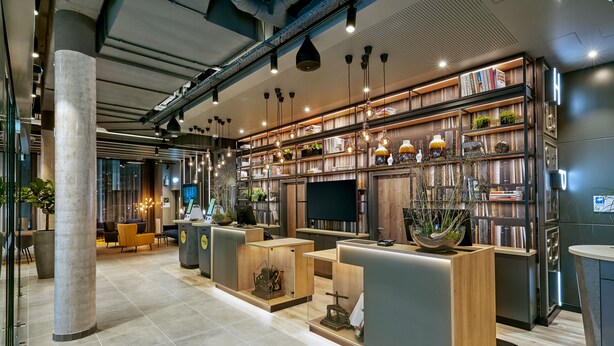 A modern hotel lobby with a reception desk, seating area, and a bookshelf filled with books and decorative items. - H2 Hotel Mainz (Mainz)
