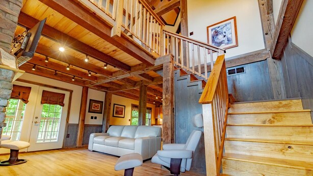 A spacious living area with a wooden staircase, a white leather sofa, and a high ceiling with exposed beams. - Panoramic Point Cabin - Beautiful Mountain Retreat for Families (Ohiopyle)
