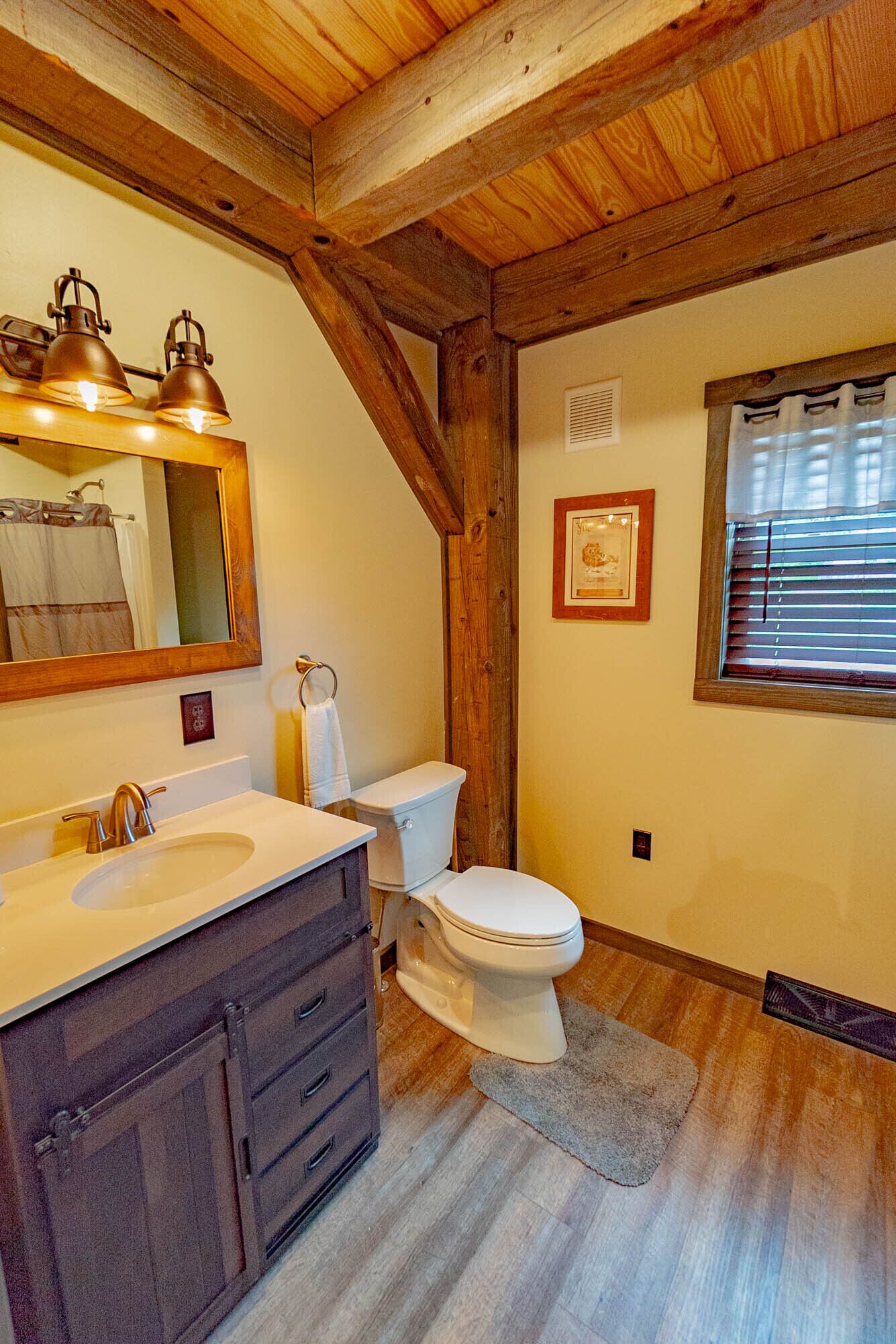 Une salle de bain avec un plafond en bois, un lavabo avec un miroir, des toilettes et une fenêtre avec des stores.