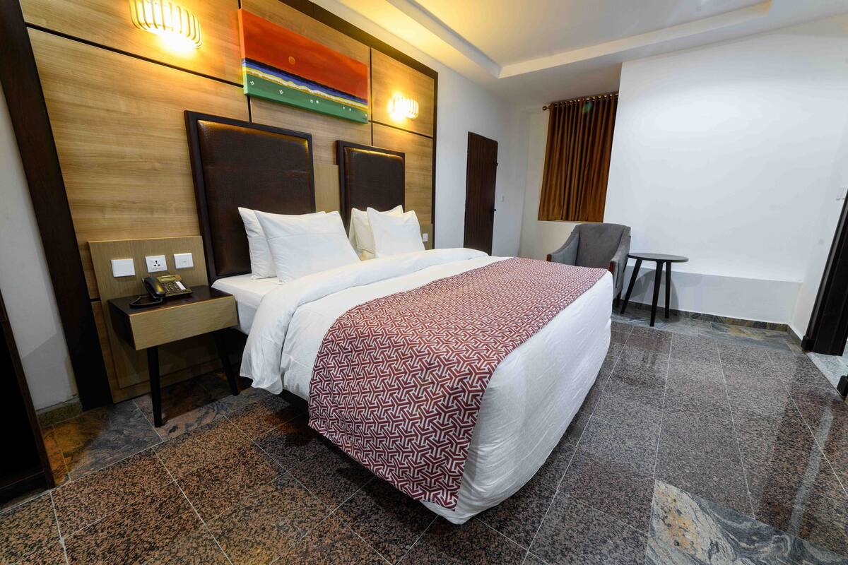 A hotel room with a large bed, a bedside table with a phone, a chair, and a small table.