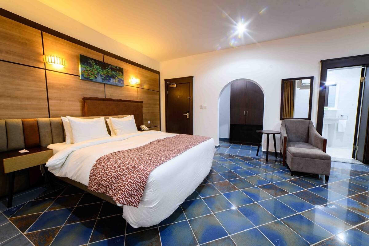 A hotel room with a large bed, a chair, a small table, and a door leading to a balcony.