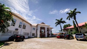 A building with a paved driveway, parked cars, and palm trees. - Alpha-Plus Apartments 91 Utako (Abuja)