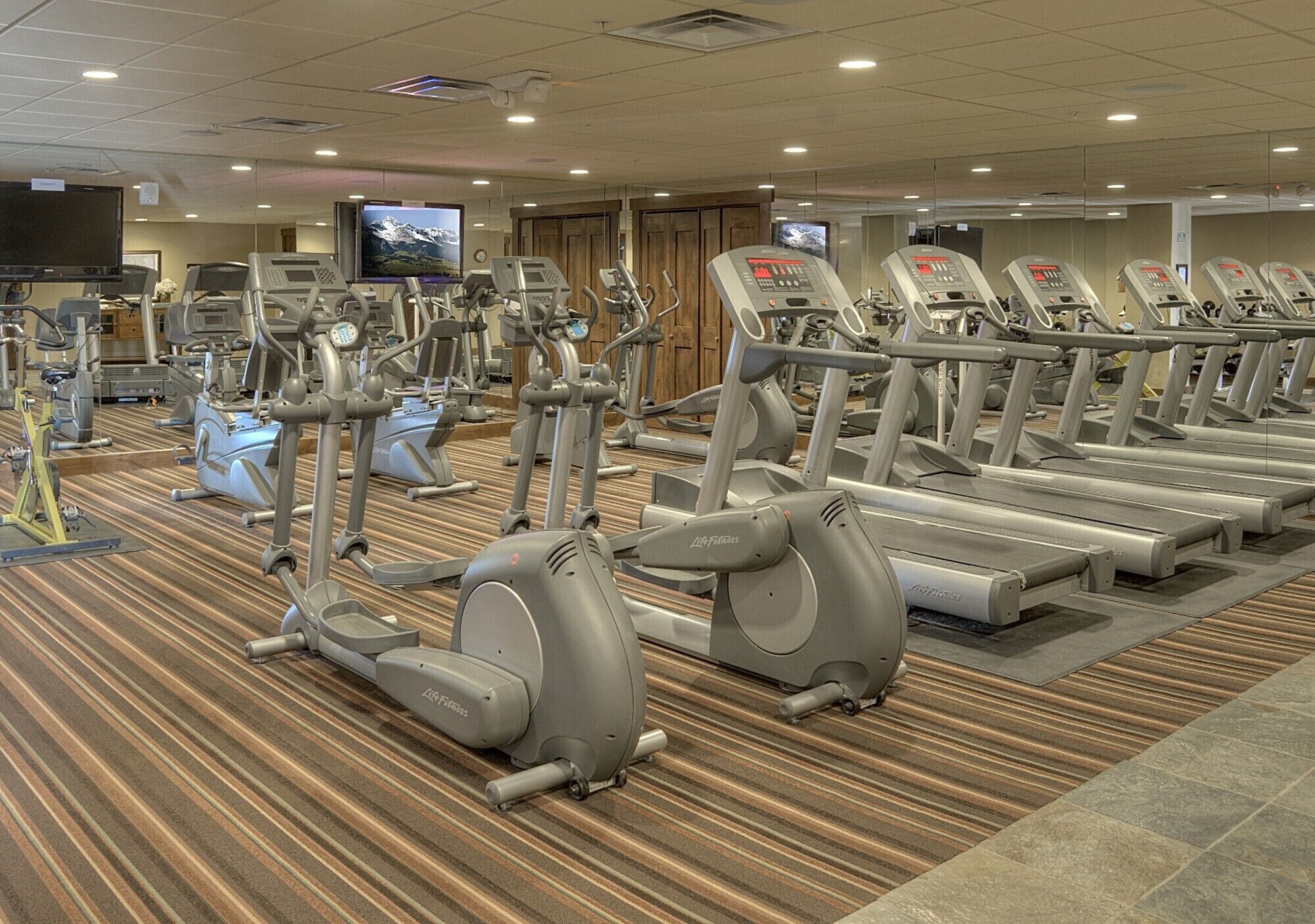 Un gimnasio con varias cintas de correr y elípticas, un televisor y piso alfombrado.