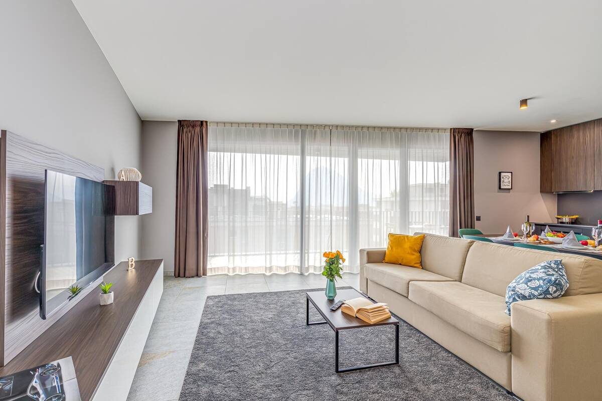 A modern living room with a flat-screen TV, a sofa, a coffee table, and a dining area with a table set for two.