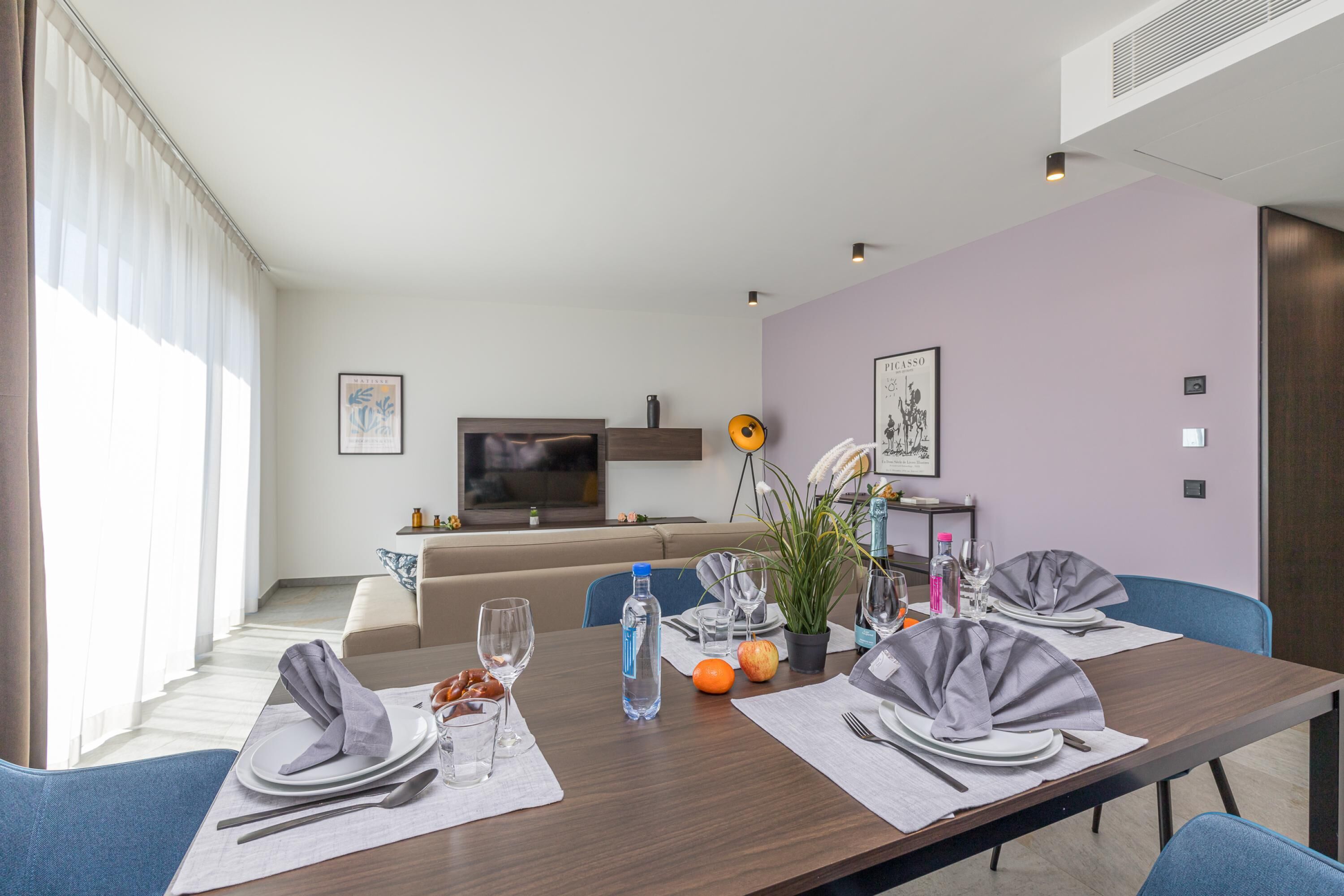 Een moderne eetruimte met een lange tafel voor vier personen, een bank op de achtergrond en een televisie aan de muur.