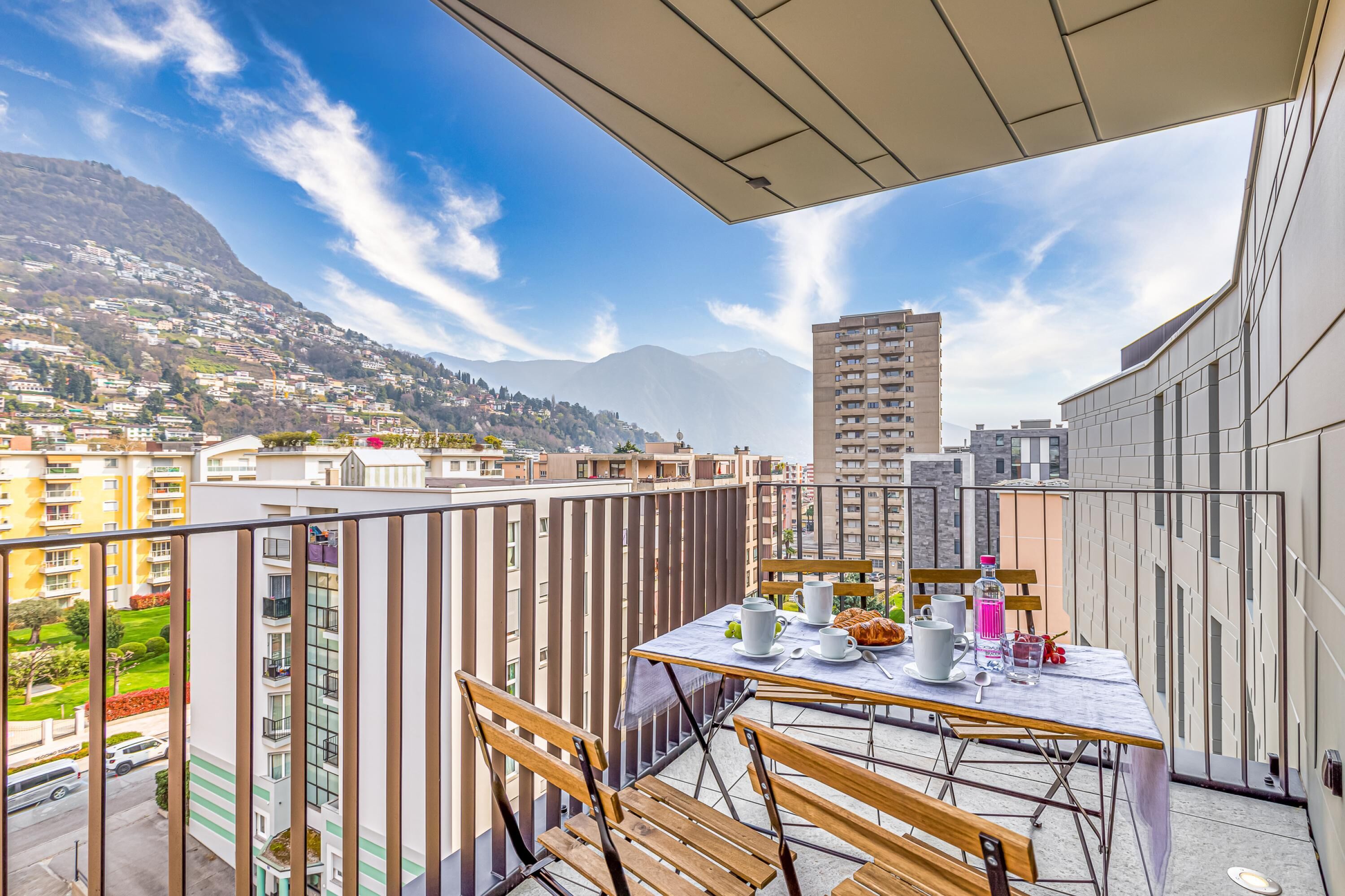 A balcony with a table set for two, overlooking a cityscape with mountains in the distance.
