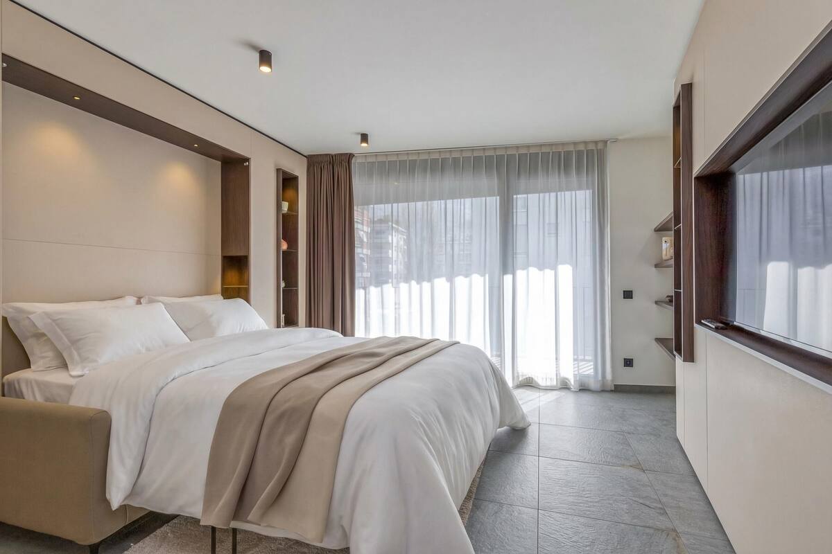 A modern bedroom with a large bed, a bedside table, a wardrobe, and a large window with curtains.