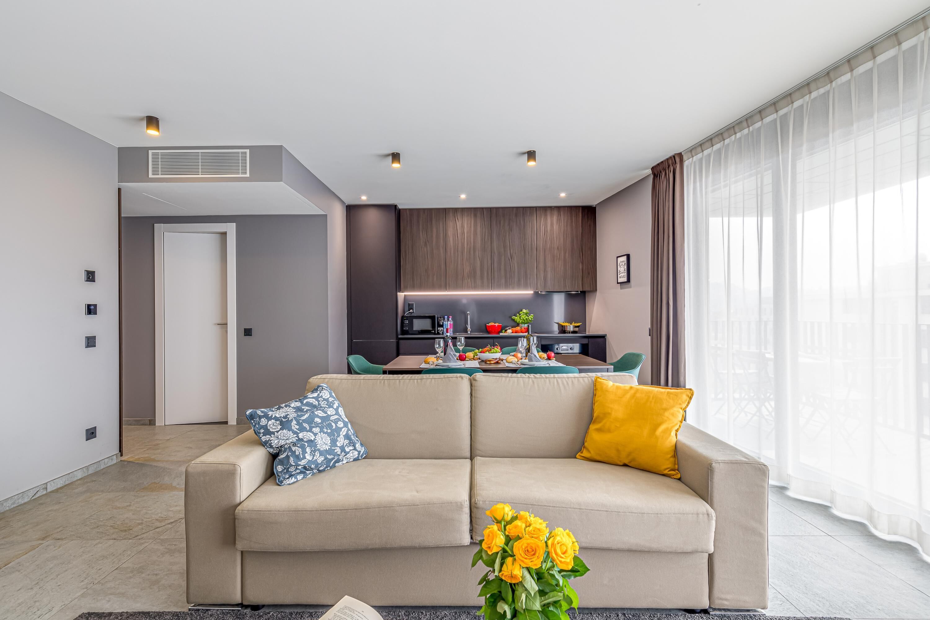 A modern living room with a sofa, a coffee table with flowers, and a kitchen in the background.