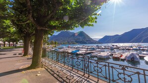 A marina with boats docked, a tree-lined walkway, and mountains in the background. - ICON H Lugano – Luxury Suites & Apartments (Lugano)