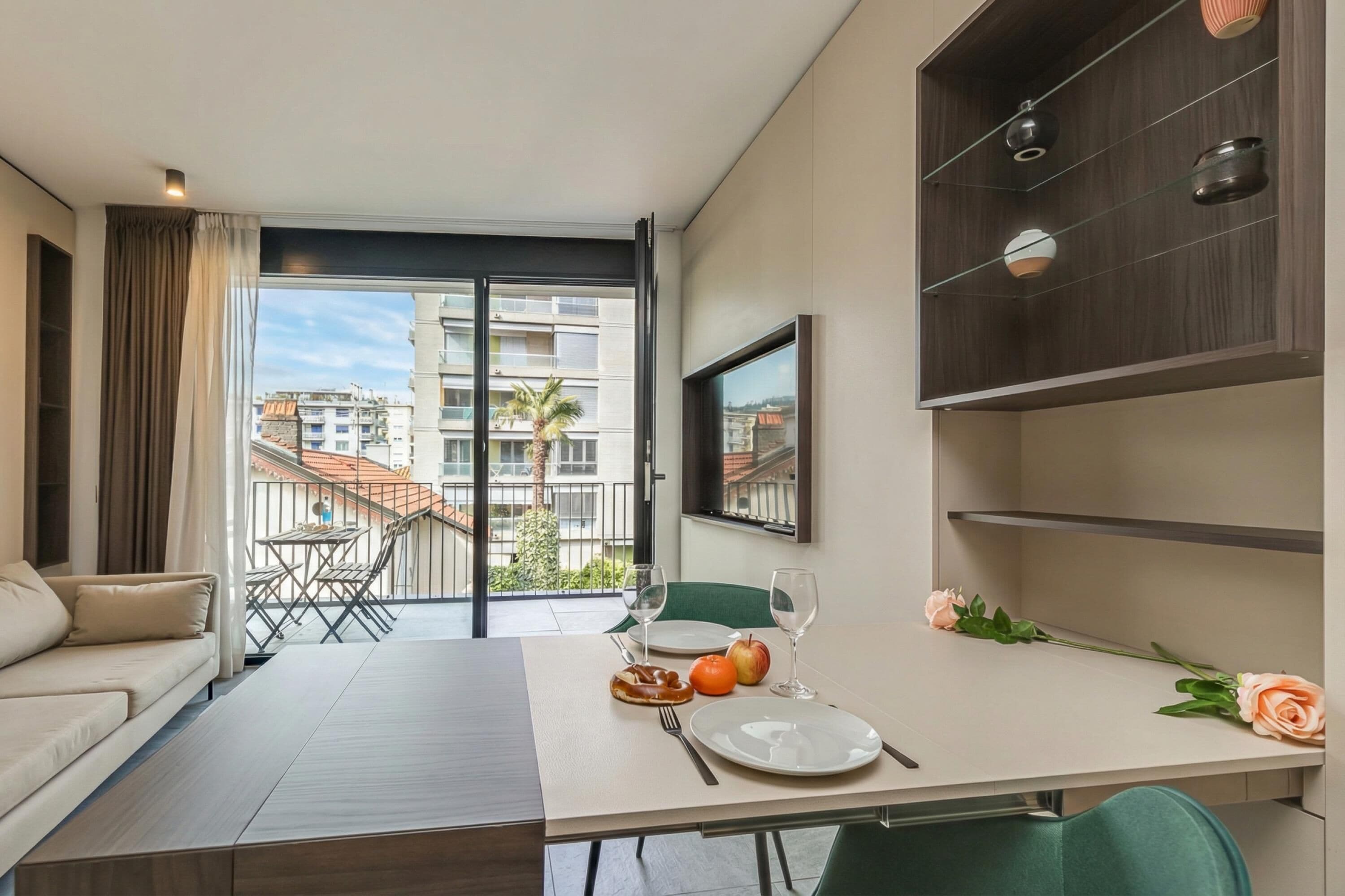 A modern dining area with a table set for two, a sofa, and a balcony with a view of buildings.