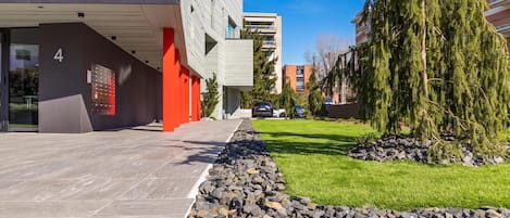 Een modern gebouw met een pad van stenen dat leidt naar een rode deur, omgeven door groen en een heldere lucht.