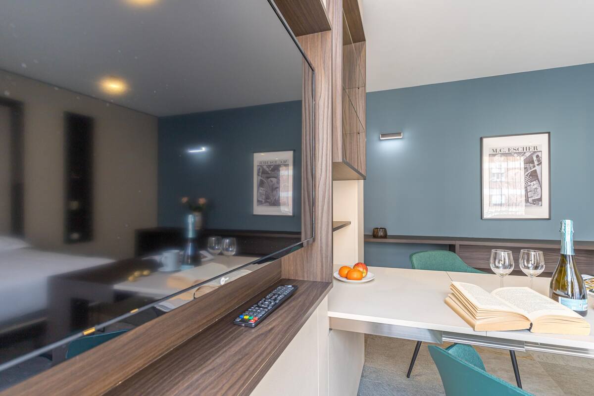 A hotel room with a bed, a desk with a remote control, a plate of oranges, two wine glasses, and a bottle of wine.