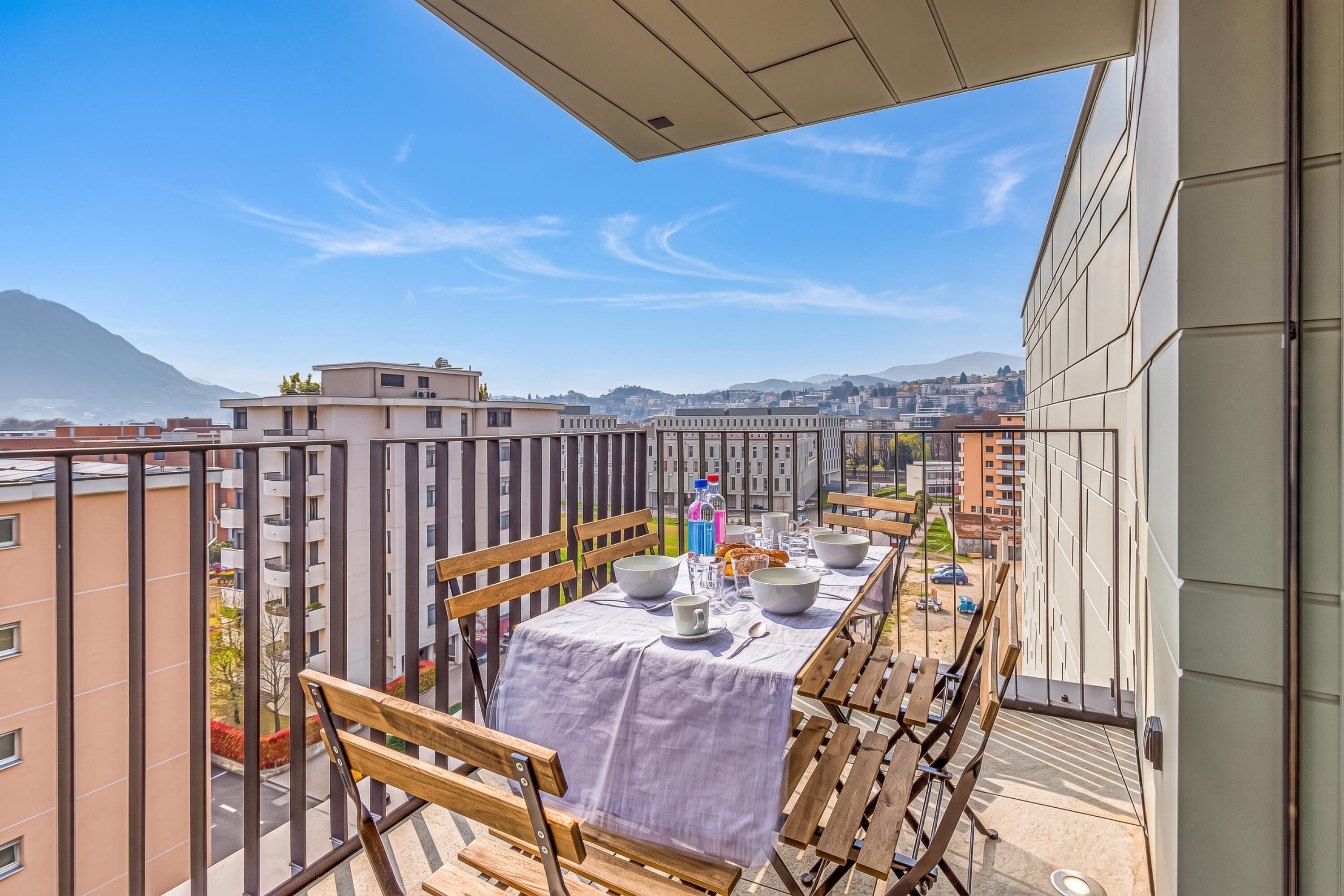 A balcony with a table set for dining, overlooking a cityscape with buildings and a mountain.