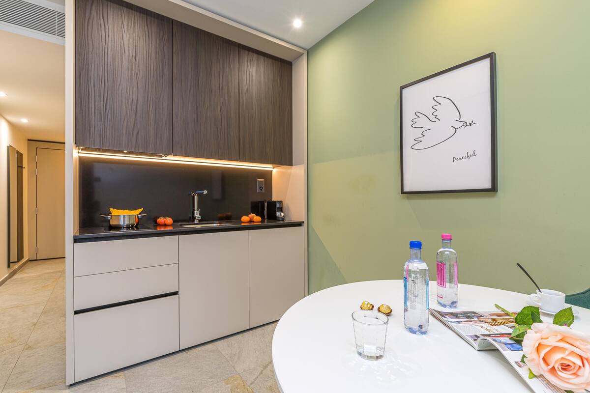 A modern kitchen with a built-in oven, a coffee machine, and a sink. There is a round table with a magazine, a glass of water, and two bottles.