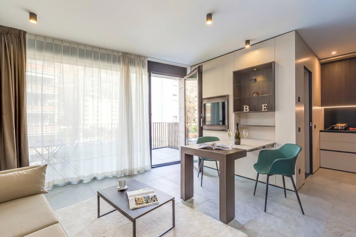 A modern living room with a sofa, coffee table, and a dining area with chairs. There is a sliding door leading to a balcony.
