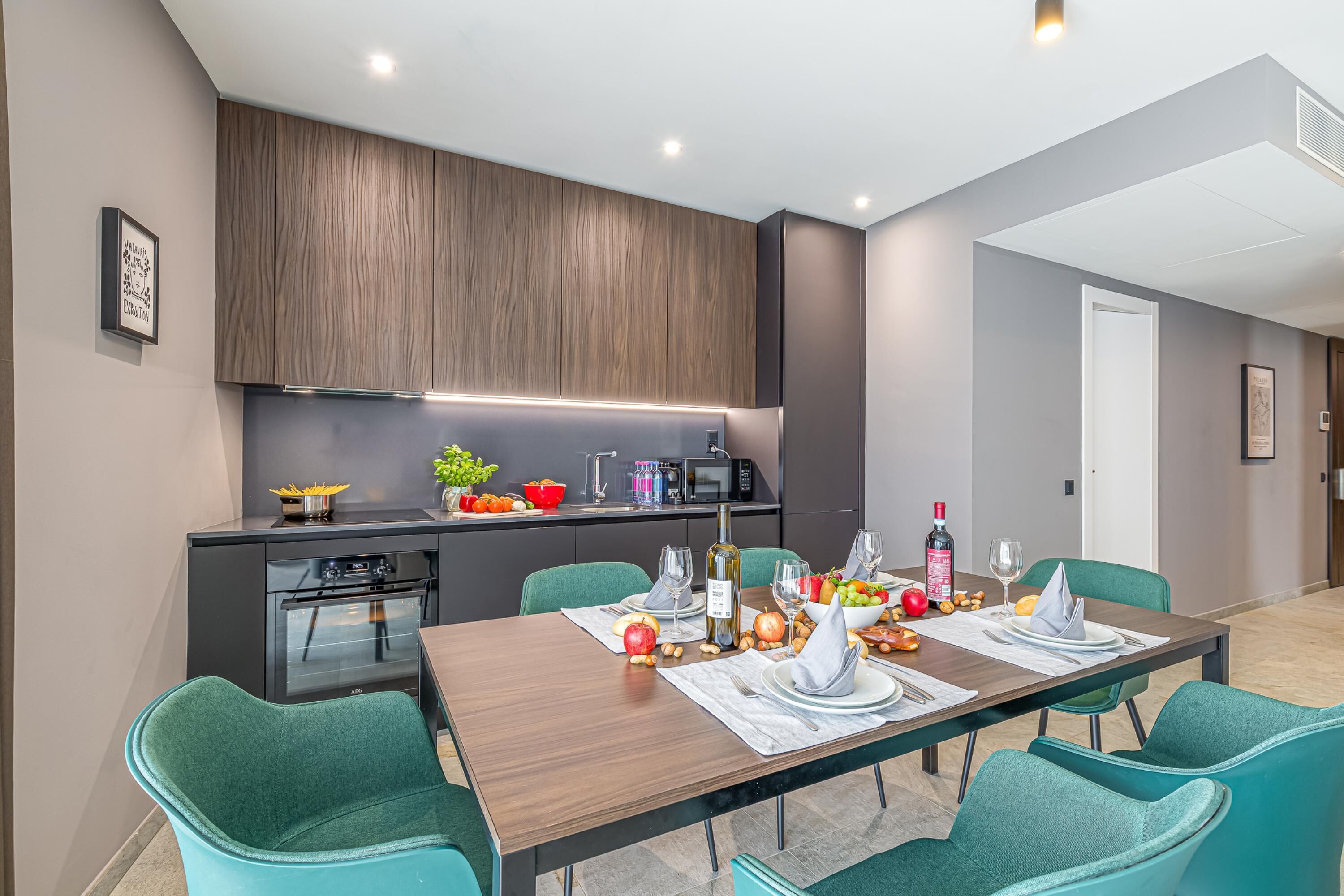 Una cocina moderna con armarios de madera oscura, un microondas integrado y un comedor con sillas color turquesa y una mesa de madera dispuesta para una comida.