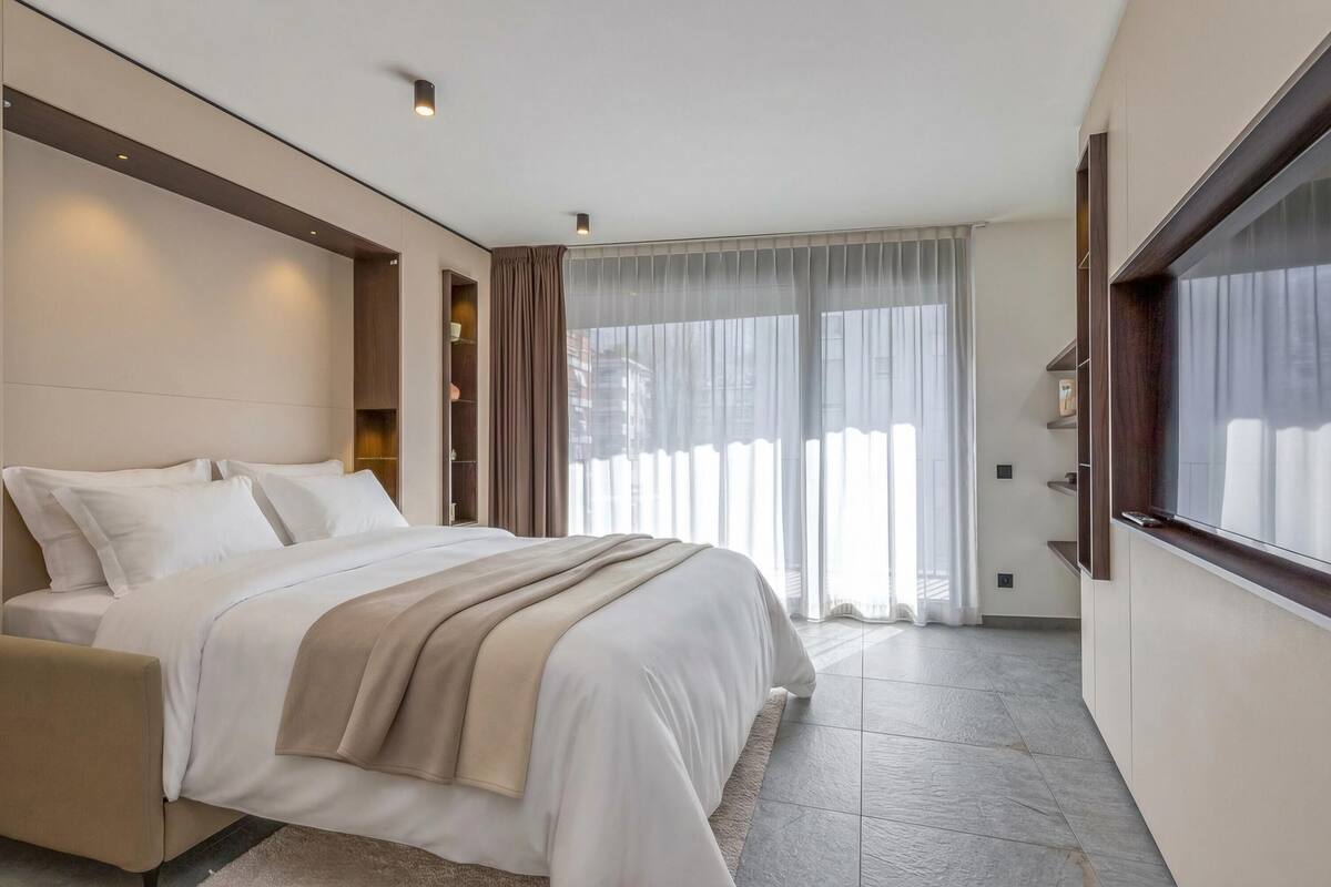 A modern hotel room with a large bed, a sofa, a built-in TV, and a large window with curtains.