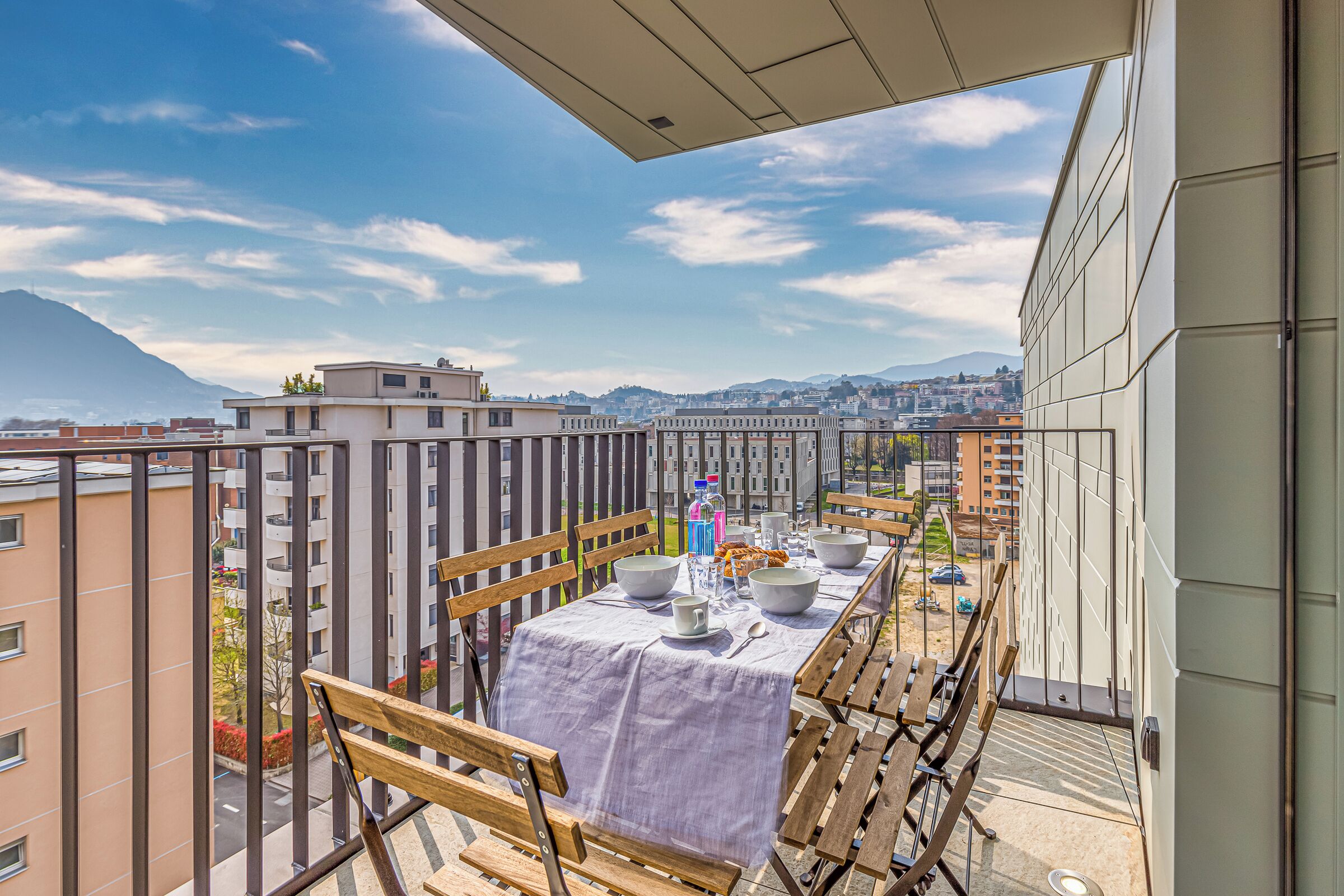 A balcony with a table set for dining, overlooking a cityscape and mountains.