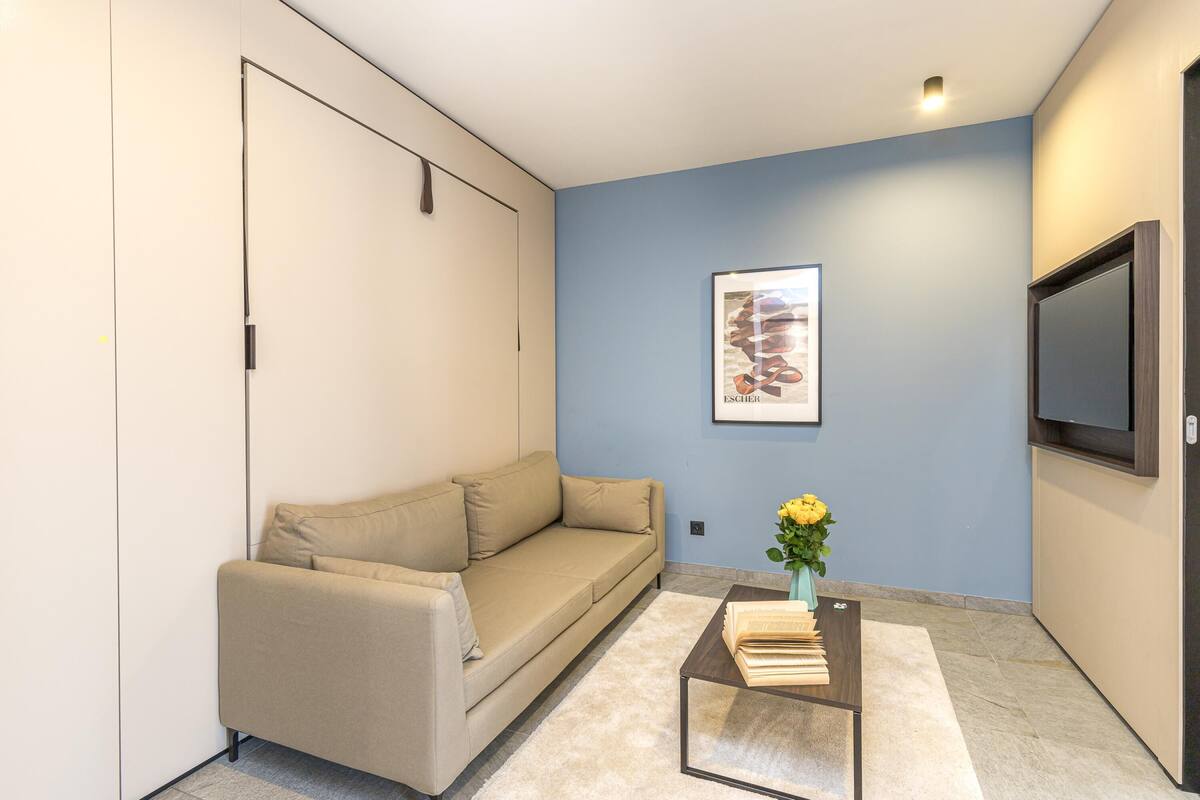 A modern living room with a beige sofa, a coffee table with books, a vase with yellow flowers, and a framed picture on the wall.