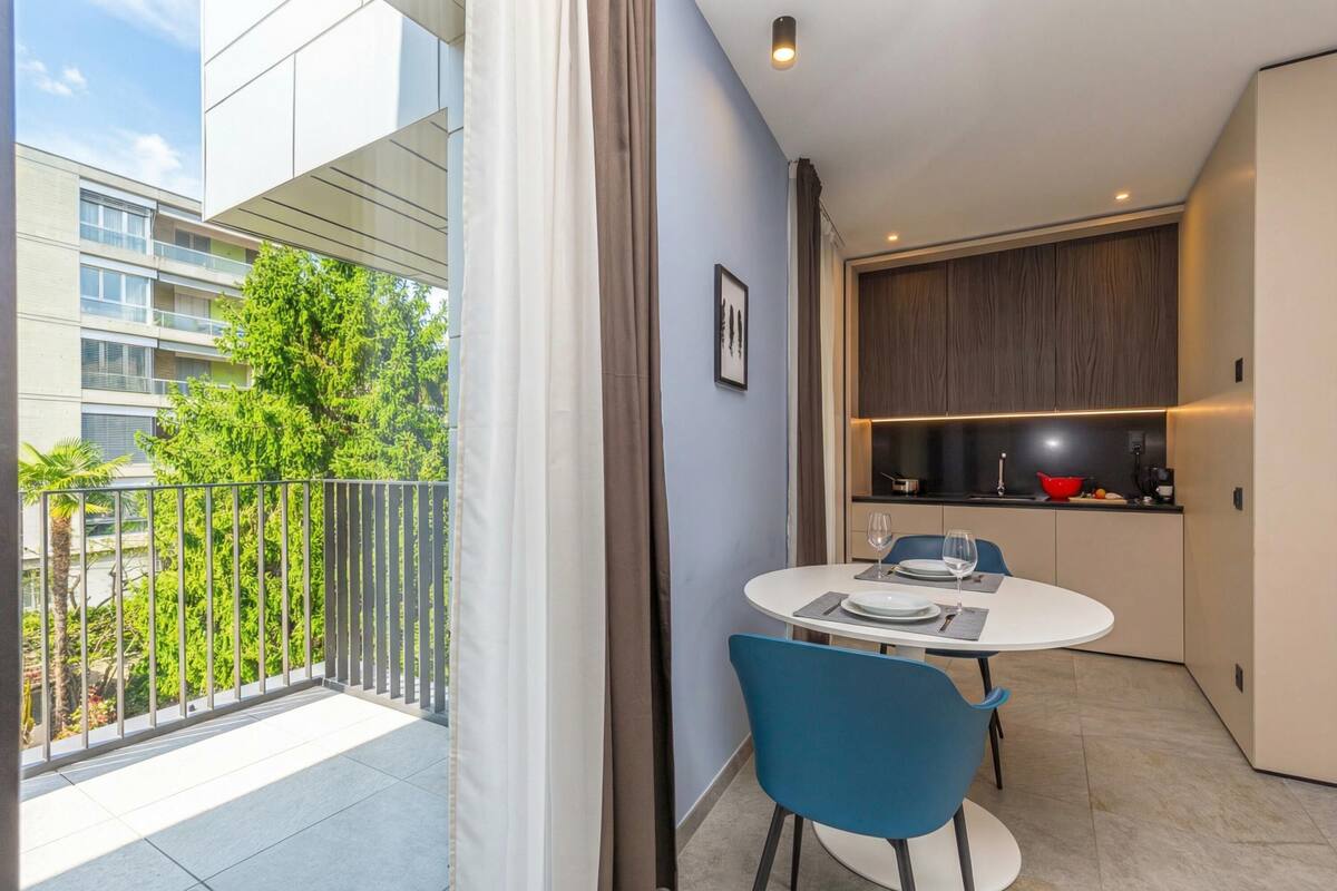 A modern apartment with a balcony, a dining area with blue chairs, and a kitchen with dark cabinets.