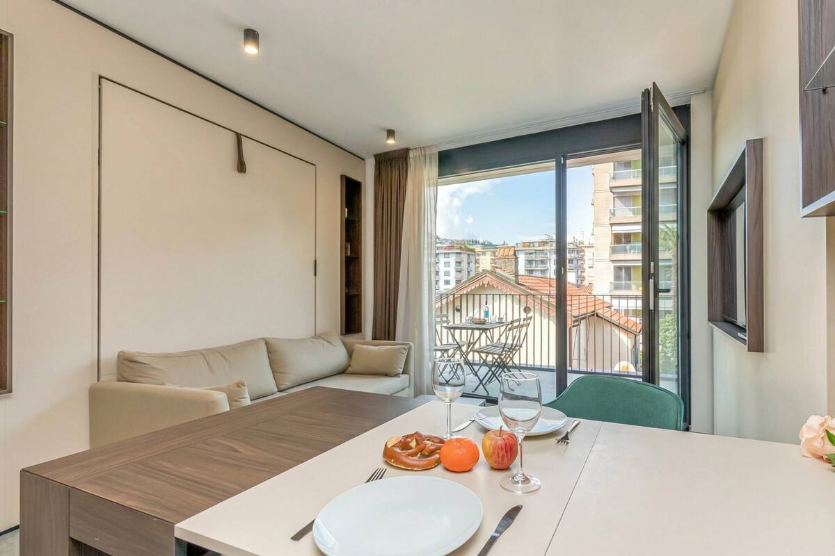 A modern hotel room with a dining area, a sofa, and a balcony with a view of buildings.