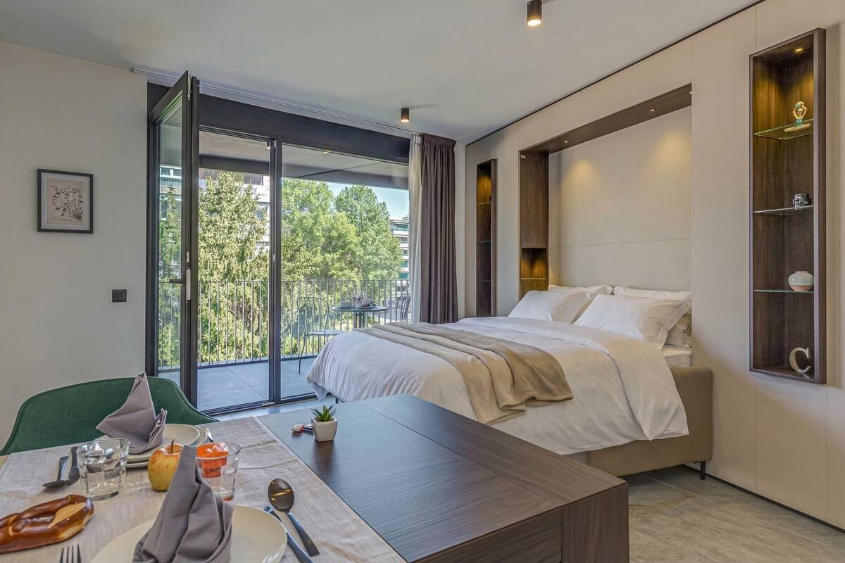 A modern hotel room with a bed, a dining table with breakfast items, a green chair, and a view of trees through a large window.