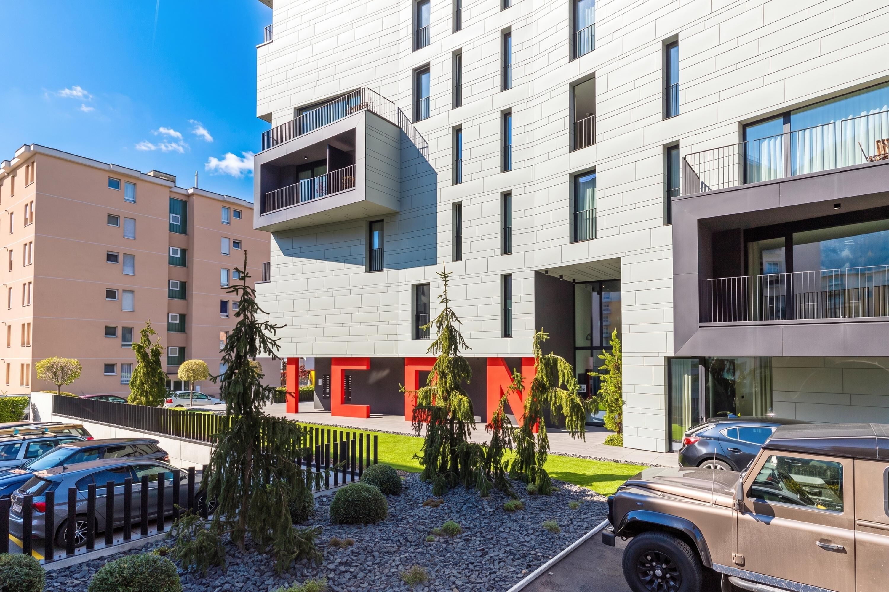 A modern building with multiple balconies, a parking area, and a small garden with trees.