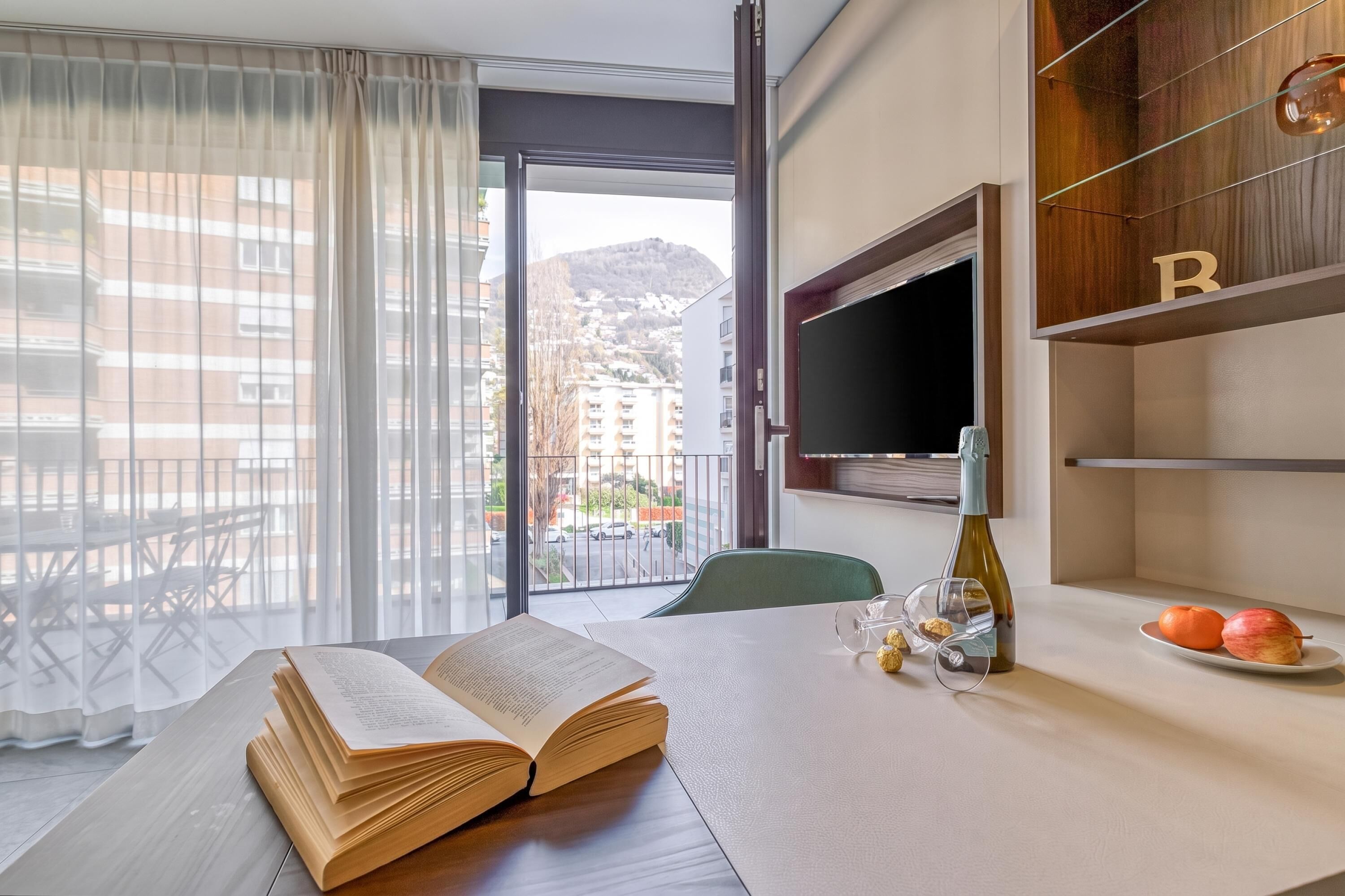 Habitación de hotel con un ventanal grande, una mesa de comedor con un libro abierto, una botella y un plato con fruta.