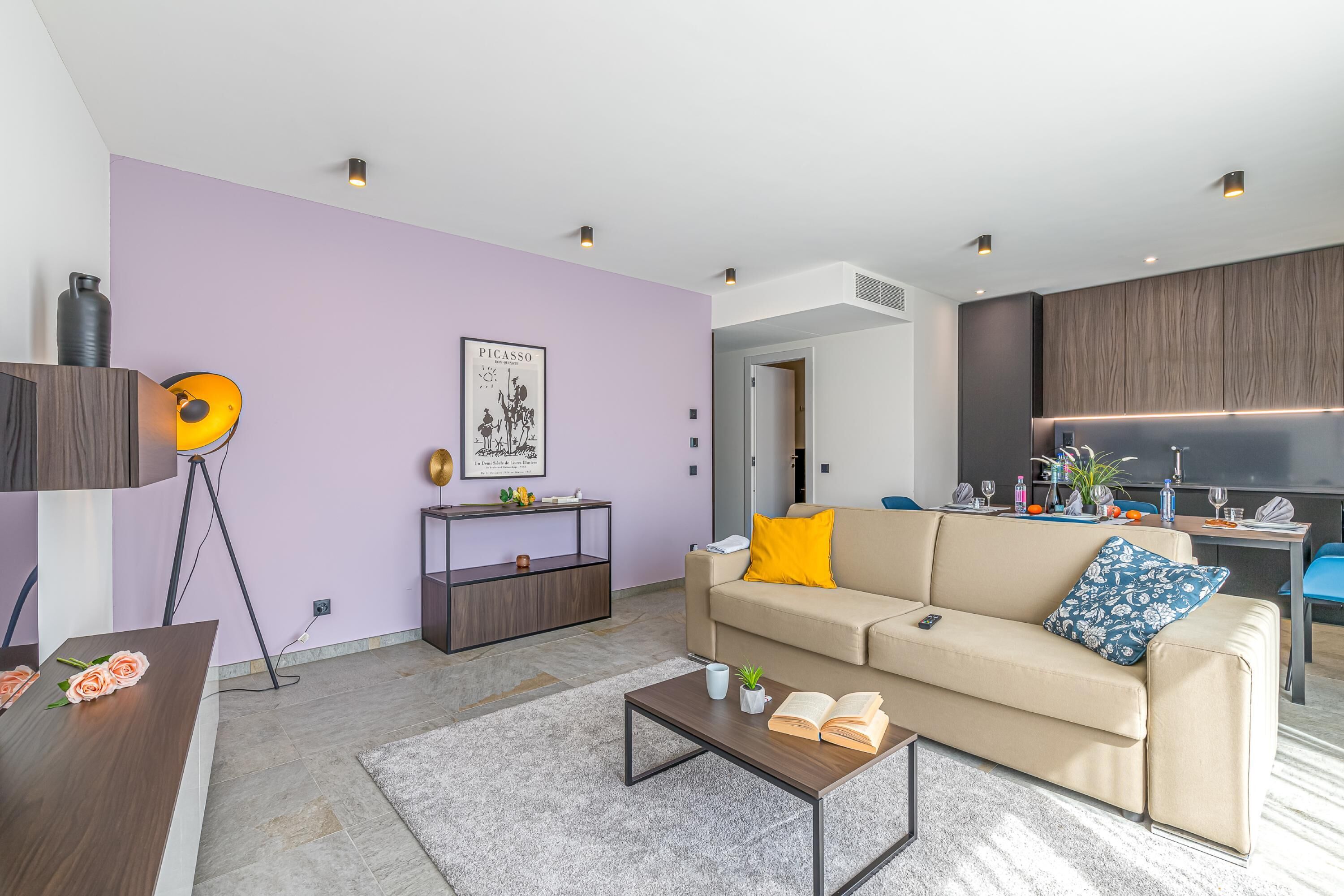 A modern living room with a sofa, coffee table, and a dining area with a kitchen in the background.
