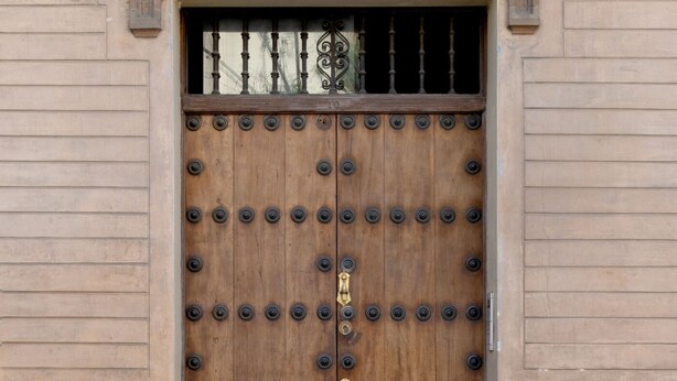 Una puerta de madera con remaches metálicos y un tirador de latón, en un edificio de ladrillo.