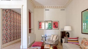 Una sala de estar con un sofá, un banco, una mesa de centro y una estantería. Hay tres obras de arte enmarcadas en la pared y una lámpara sobre la mesita auxiliar.