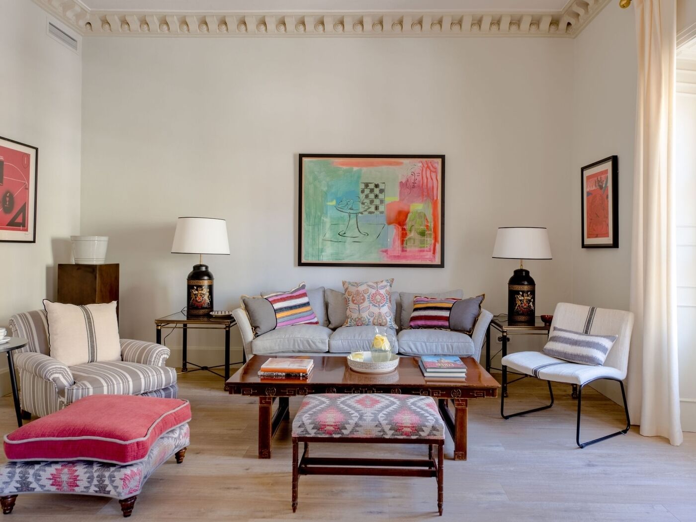 Ein Wohnzimmer mit einer Couch, Sesseln, einem Couchtisch und einem gemusterten Pouf. Zwei Beistelltische mit Lampen und ein großes abstraktes Gemälde an der Wand.