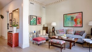 Una sala de estar con un sofá, sillones, una mesa de centro y una mesita auxiliar. Hay cuadros enmarcados en la pared y una lámpara sobre la mesita.